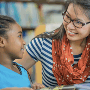 Applied Behavior Analysis Technician (ABAT) instructor smiling and engaging with a young student during a reading activity in a classroom or library setting.